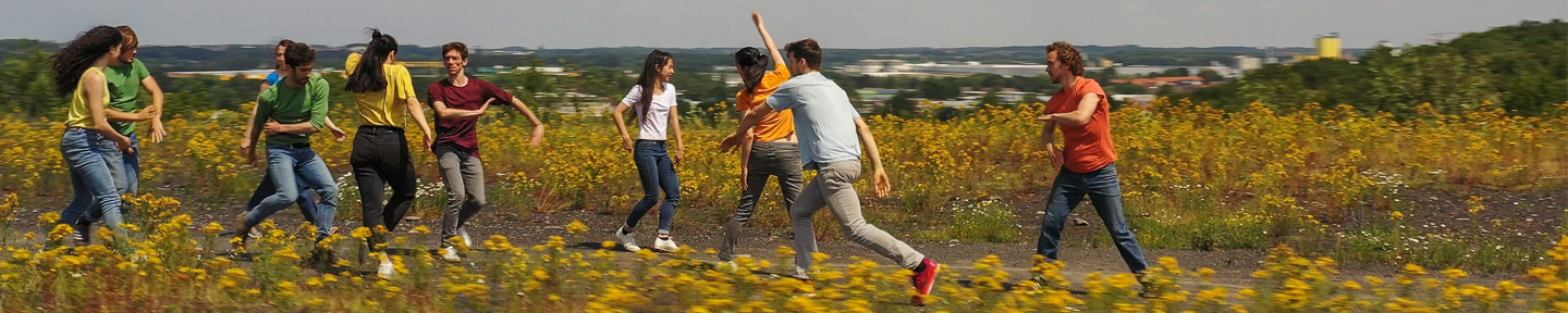 Mehrere Personen in urbaner Kleidung tanzend auf einer Wiese mit gelben Blumen. Im Hintergrund verschwommen das Panorama einer Stadt