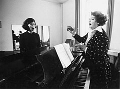 In schwarz-weiß: Prof. Rita Streich singend vor einem Piano. Ihr gegenüber ein:e Studierende:r.
